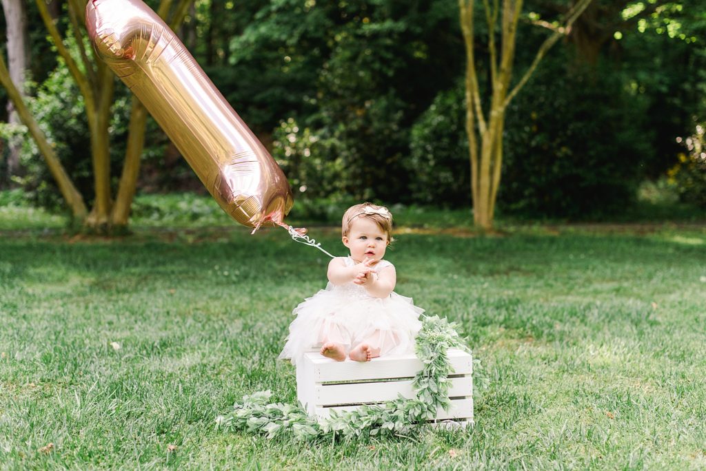 one year old girl in white dress on white wooden box holding pink #1 balloon