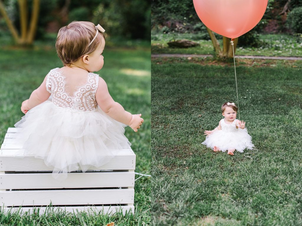 one year old girl in white dress on white wooden box holding pink balloon