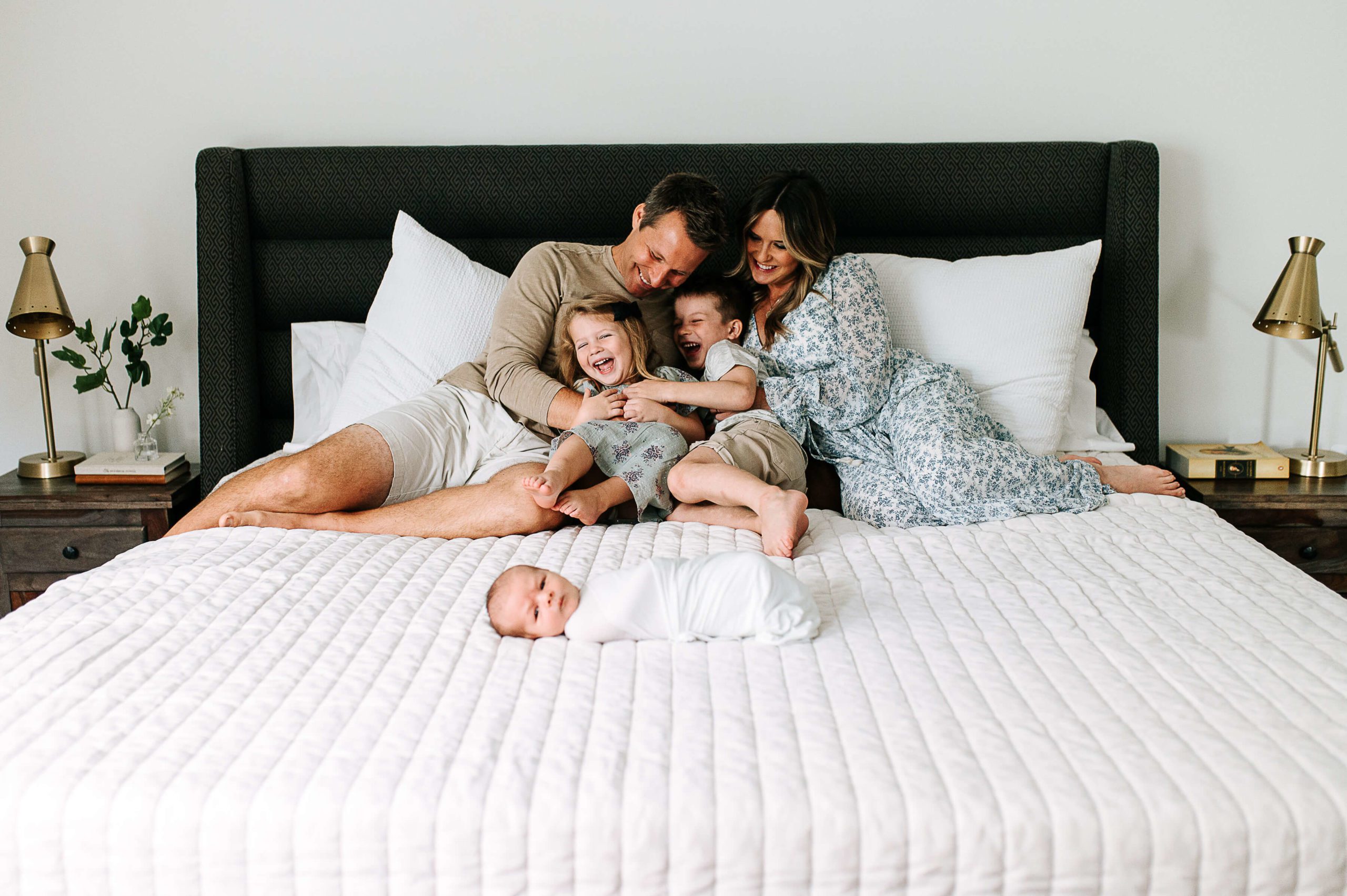 family giggles on bed with white bedding and swaddled newborn baby